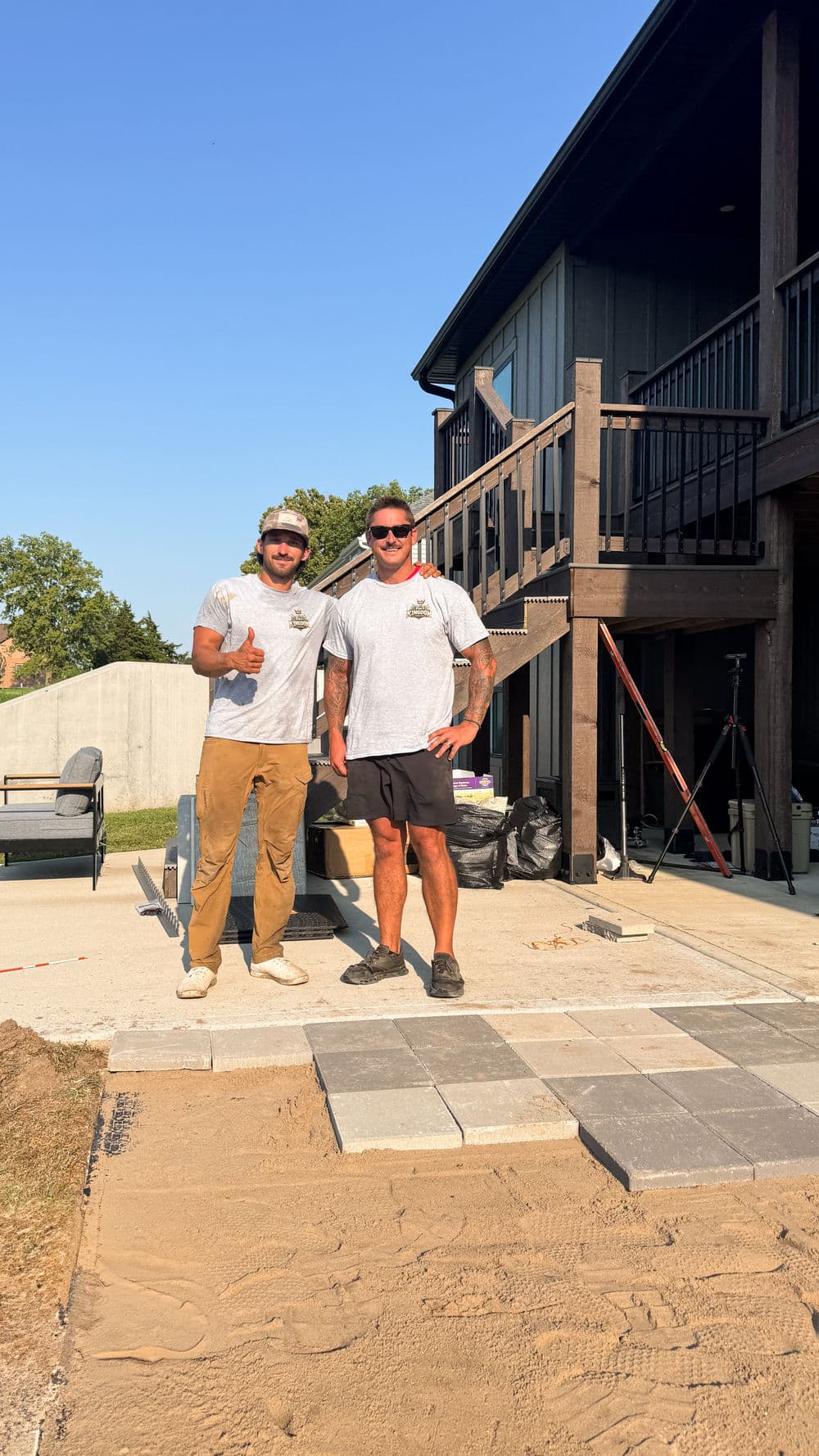 Two workers smiling on a patio installation site with pavers and a modern home in background.